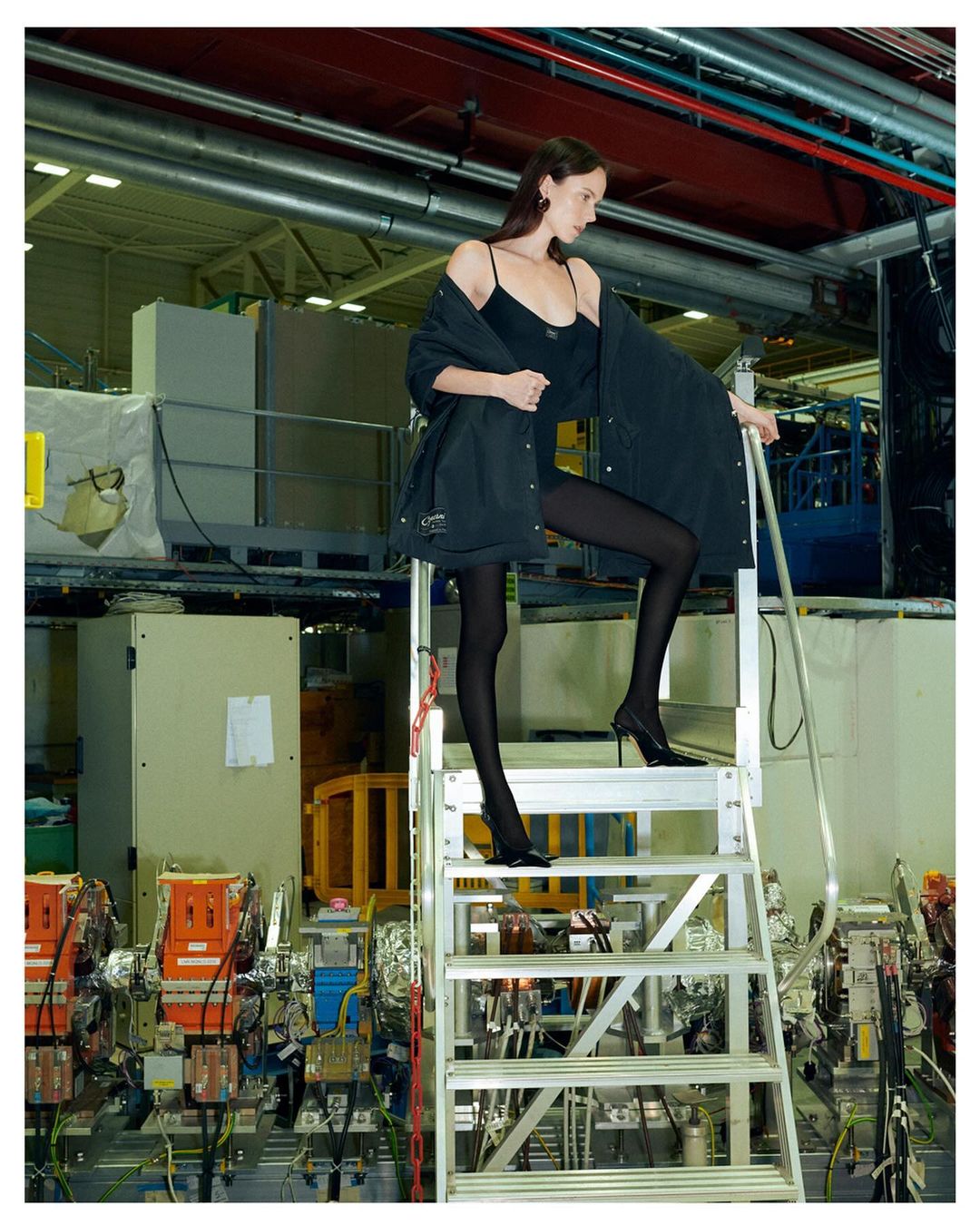 Fashion editorial shot: A slender woman poses in a black bodysuit and jacket on aluminum stairs inside an industrial facility.