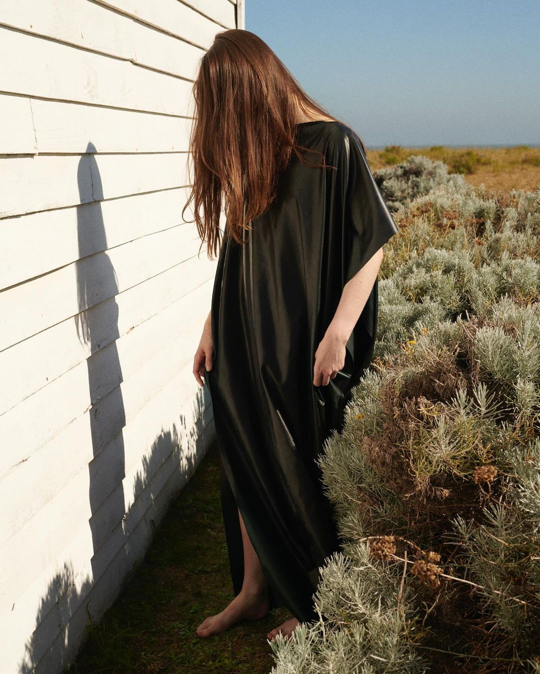 Woman with long brown hair covering her face wearing a long black dress standing beside a white wooden wall outdoors.