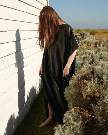 Woman with long brown hair covering her face wearing a long black dress standing beside a white wooden wall outdoors.