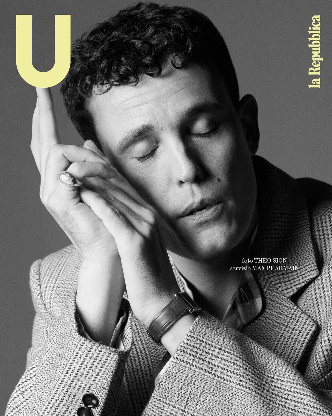 Black and white magazine cover featuring a portrait of a fair-skinned male model with curly hair, eyes closed, and hands near his face.