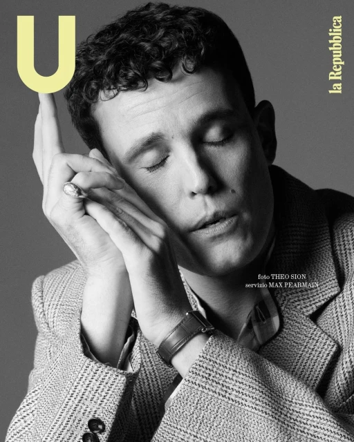 Black and white magazine cover featuring a portrait of a fair-skinned male model with curly hair, eyes closed, and hands near his face.