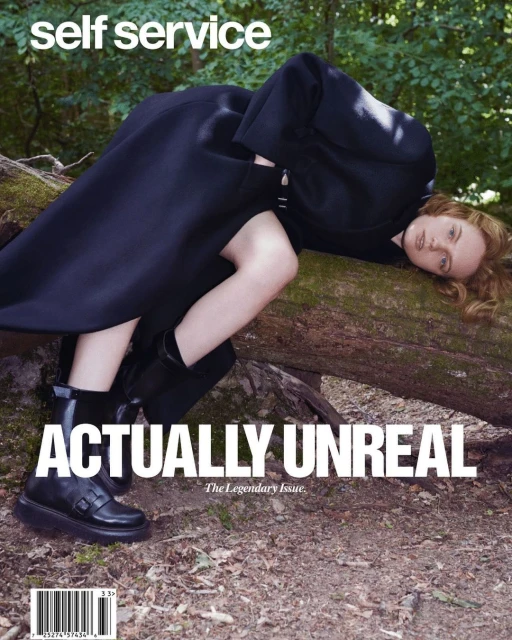 Fashion editorial magazine cover featuring a fair-skinned, red-haired woman in a black coat posing on a fallen tree in a forest setting.
