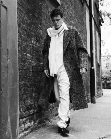 Man walking on a concrete sidewalk alongside a weathered brick wall wearing a tweed overcoat and white scarf.