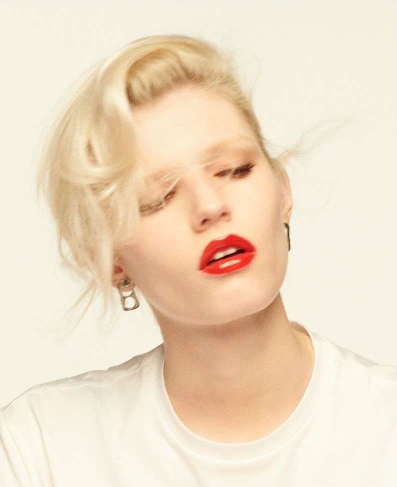 Close-up portrait of a woman with blonde hair and glossy red lipstick wearing a white t-shirt against a plain backdrop.