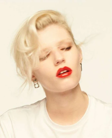Close-up portrait of a woman with blonde hair and glossy red lipstick wearing a white t-shirt against a plain backdrop.