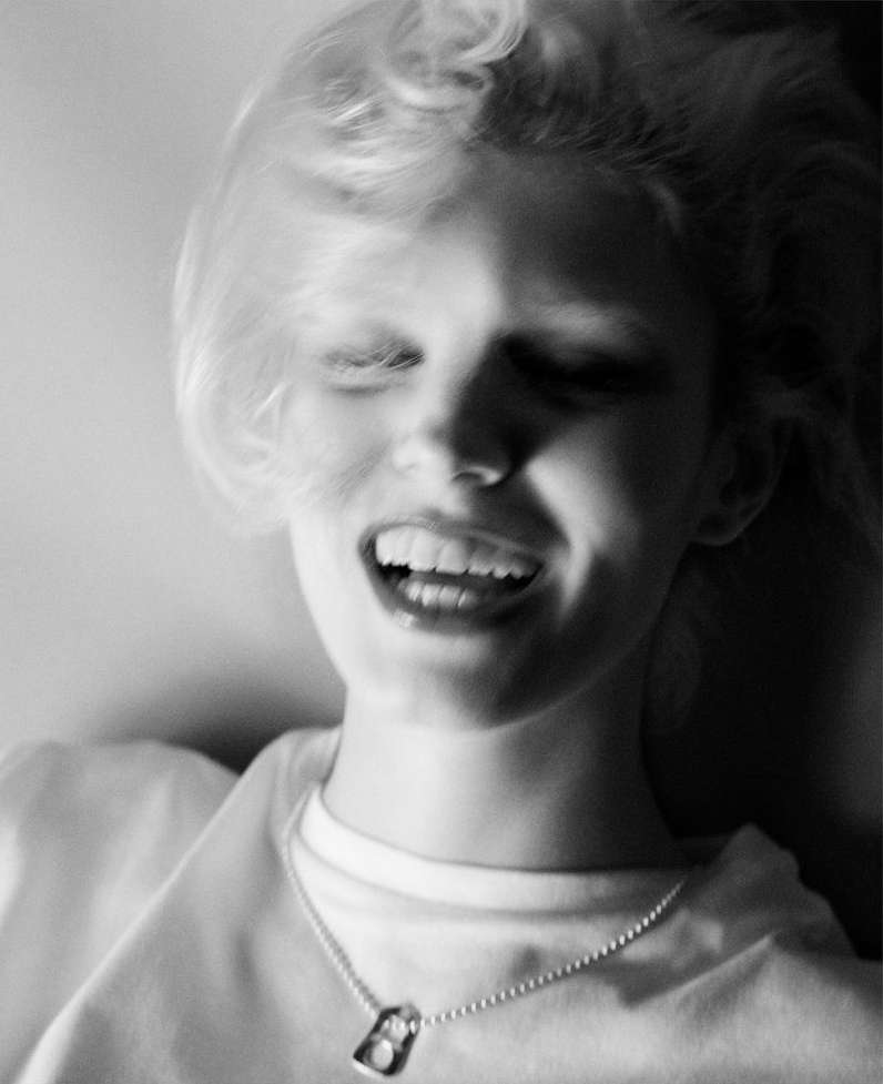 Monochrome portrait of a fair-skinned young adult woman laughing with her eyes closed, wearing a t-shirt and a dog tag necklace.