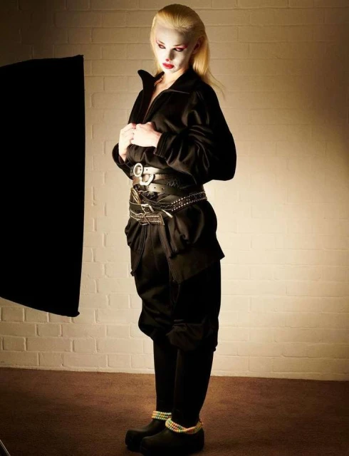 Full-shot studio portrait of a blonde adult female posing with hands on her black zip-up jacket and a decorative belt, and low lighting.