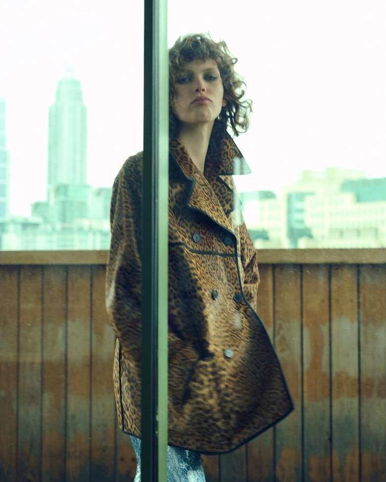 Adult female model poses in a leopard-print coat, shot in an urban setting with city buildings visible in the background.