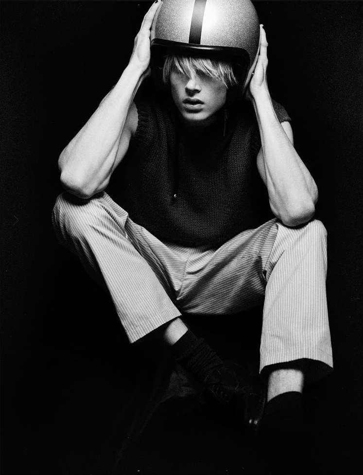 Black and white portrait of person in helmet sitting with hands holding the helmet, wearing sweater vest and striped pants, indoor.