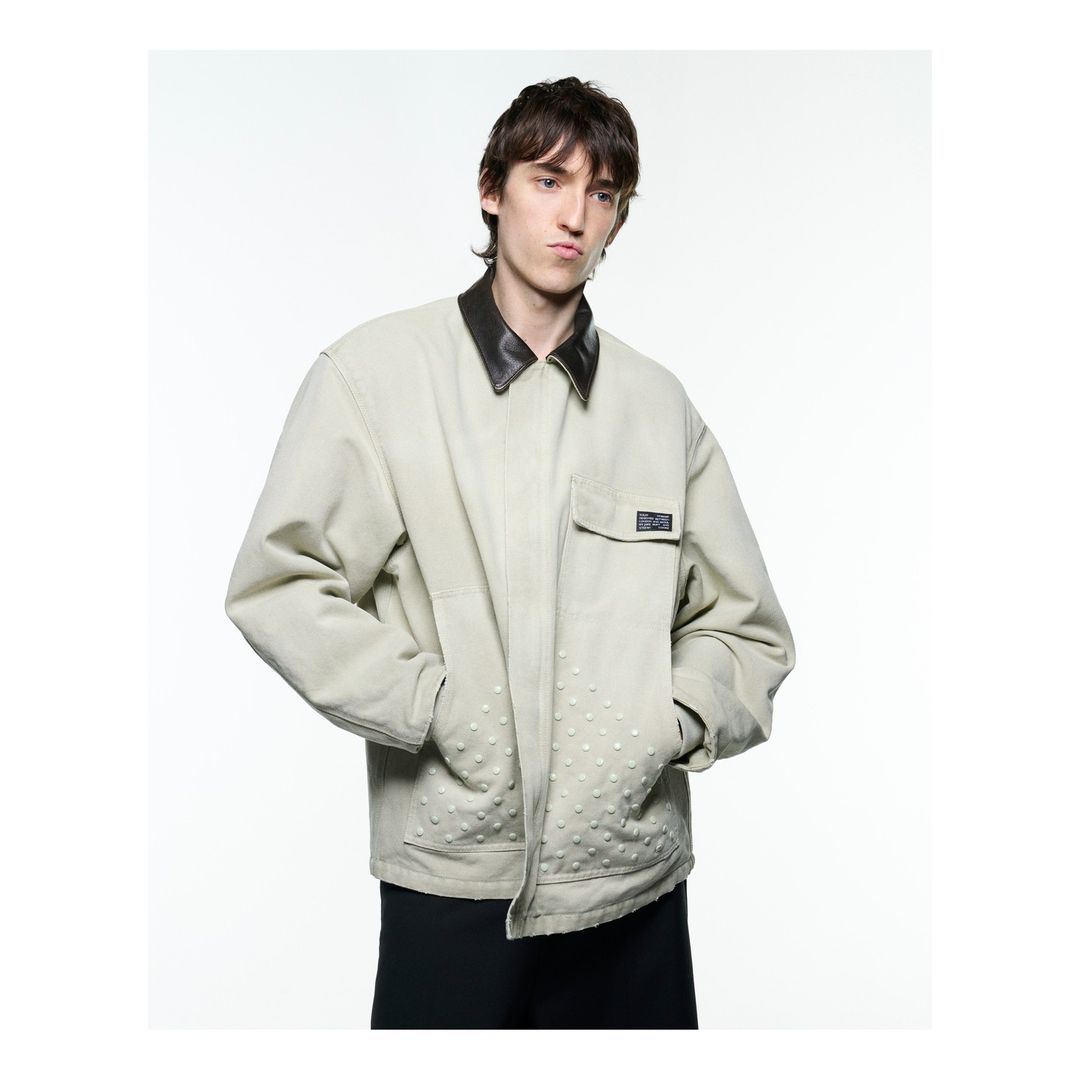 An adult male poses in a light jacket with a dark collar, against a bright white backdrop in a studio setting with a neutral expression.