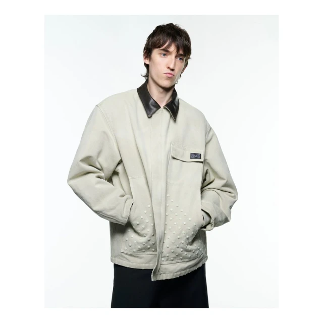 An adult male poses in a light jacket with a dark collar, against a bright white backdrop in a studio setting with a neutral expression.