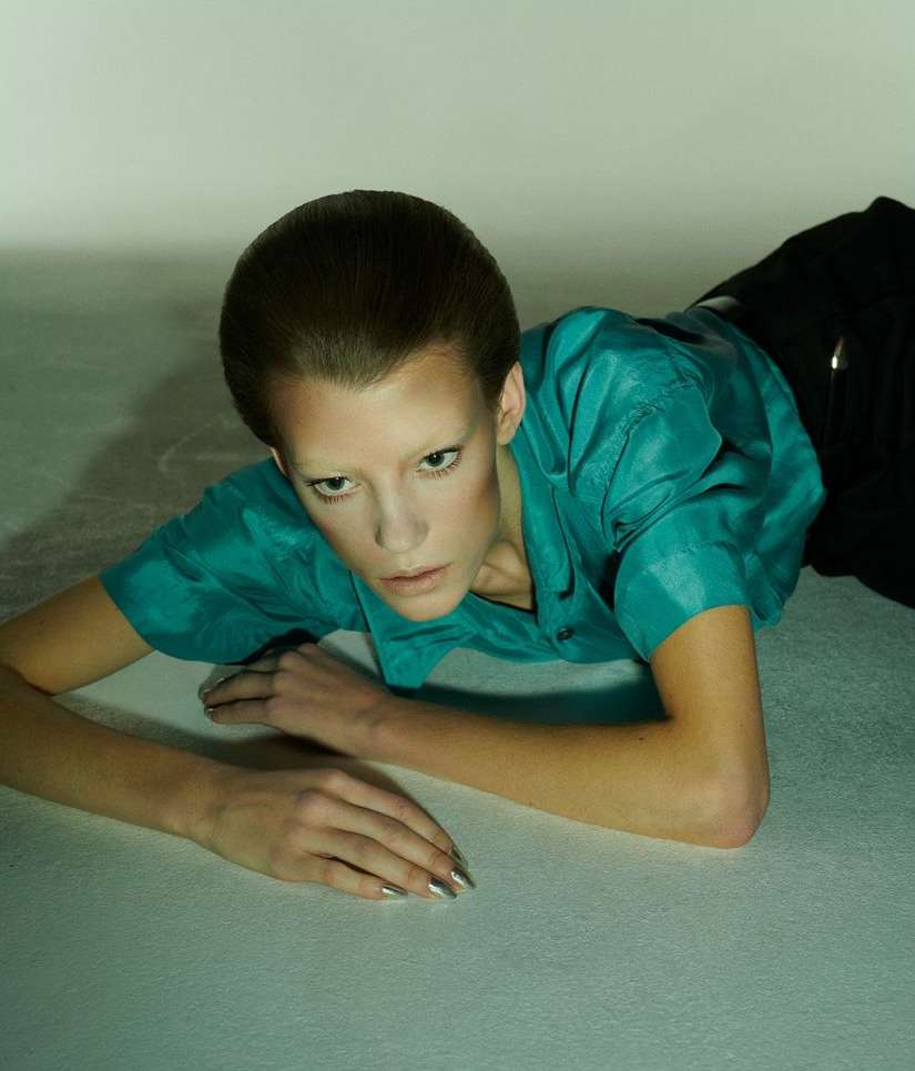 Woman with pale skin and slicked-back dark hair in a teal shirt lying on a reflective surface in a studio with even lighting.