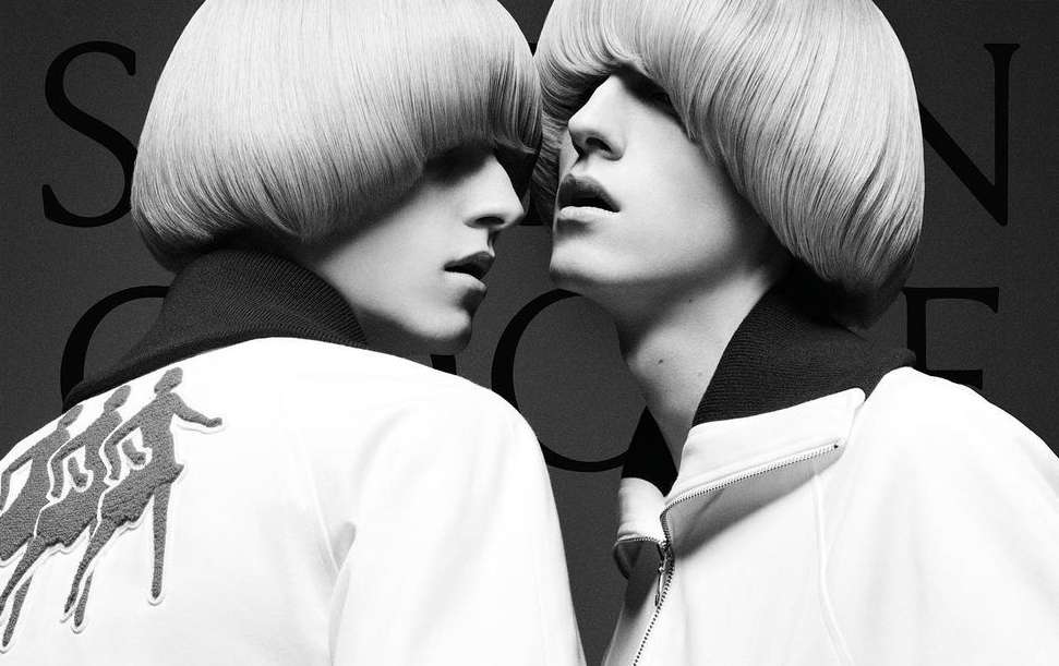 Black-and-white portrait of two individuals with matching blonde bowl cuts, wearing collared jackets with a graphic of figures on the back.