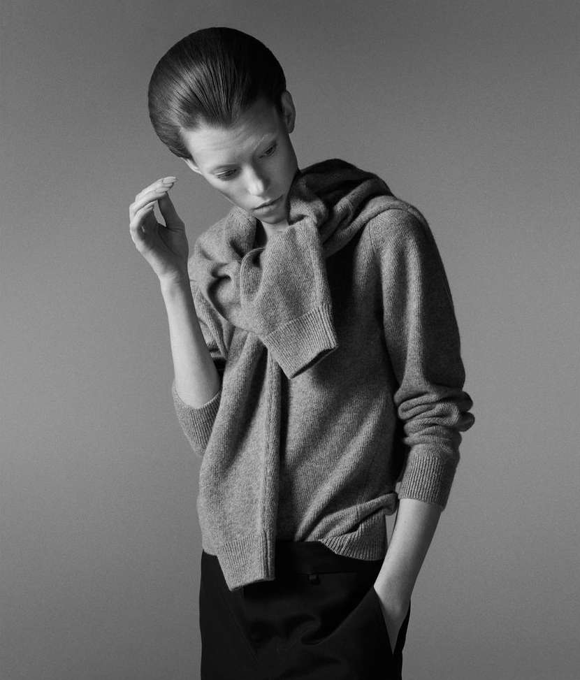 Model with slicked-back hair wearing a grey sweater over shoulders against a neutral grey studio background