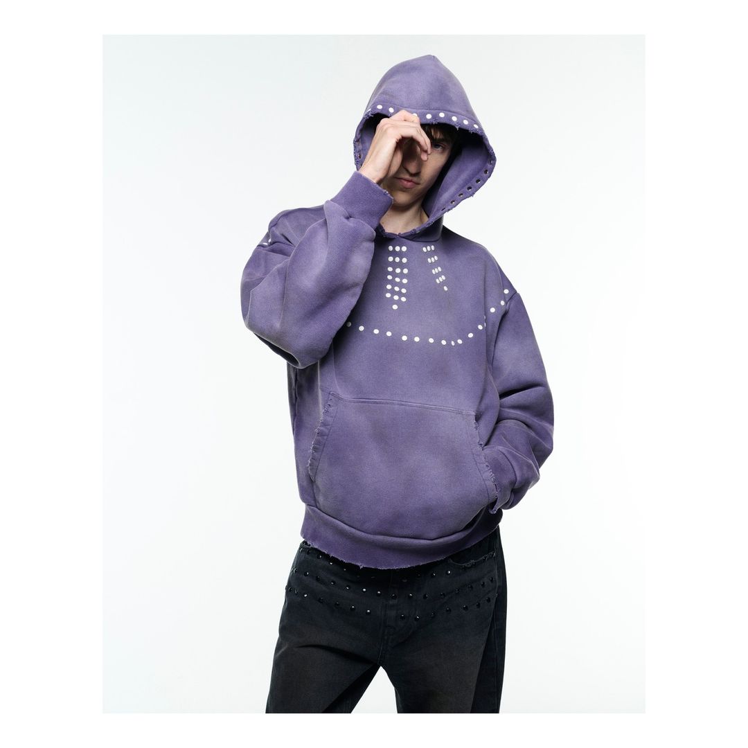 Person wearing a purple hoodie with white studs posing in a photo studio with white seamless backdrop looking towards camera.