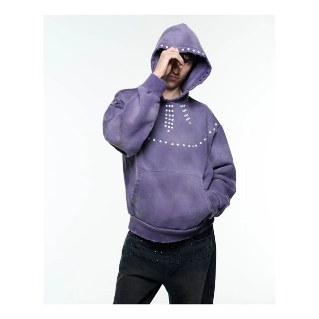 Person wearing a purple hoodie with white studs posing in a photo studio with white seamless backdrop looking towards camera.