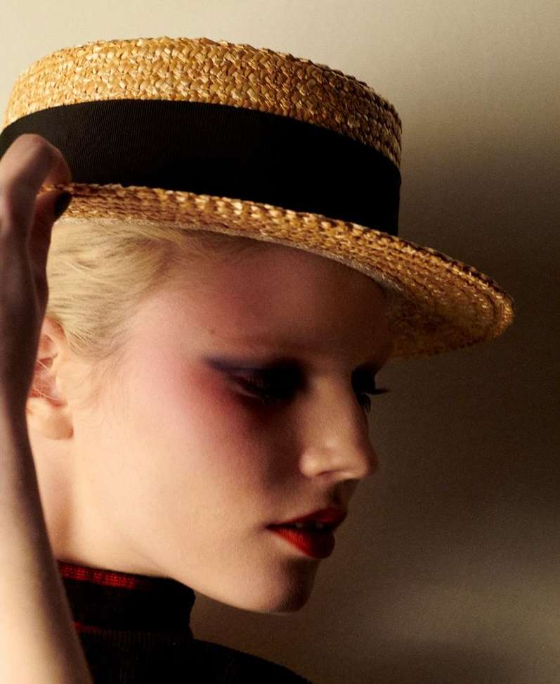 Close-up portrait of a fair-skinned adult female wearing a straw boater hat with a black band, dark eye makeup, and red lipstick.