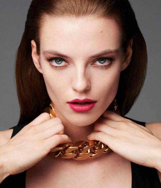 Close-up studio shot of a woman wearing a gold chain necklace, pink lipstick, with dark hair pulled back, posing against a grey background.