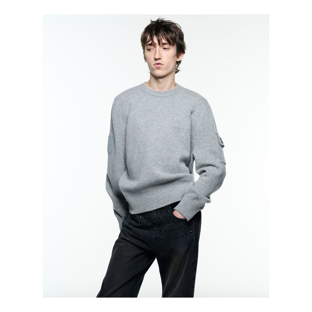 Studio shot of a young adult male wearing a grey sweater and dark pants, posing against a bright white backdrop, fashion editorial.