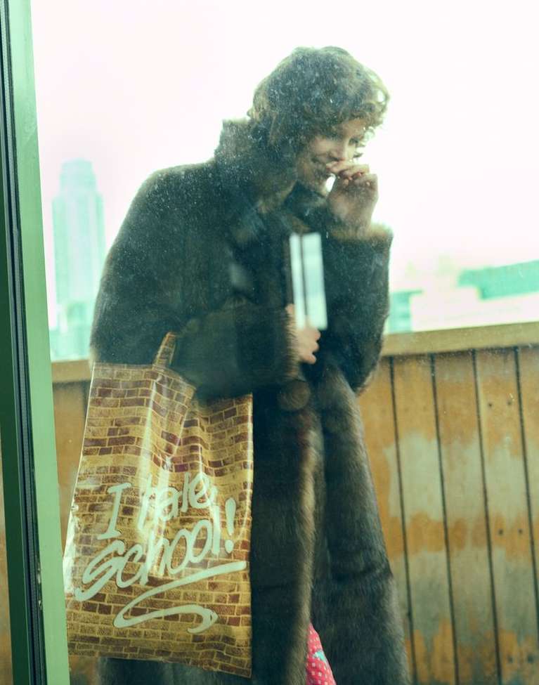 Woman in a brown fur coat and brown brick-patterned tote bag with white ‘I Hate School’ text, looking out of a window, city skyline visible.