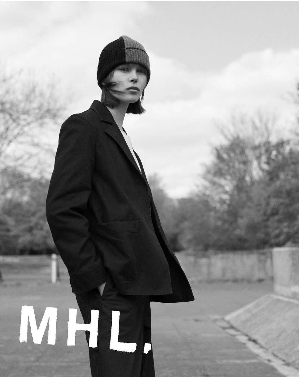 Black and white portrait of a model wearing a dark blazer and color-blocked beanie outdoors with trees in the background.