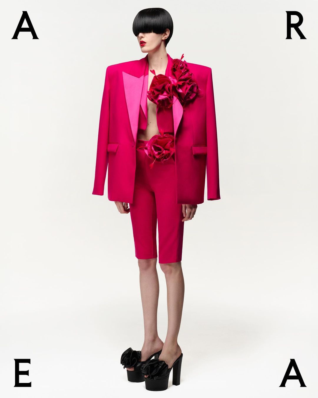 Fashion model poses in a pink suit with flower details and platform heels against a white background, studio shot.