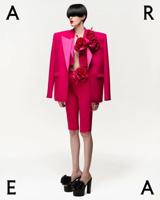 Fashion model poses in a pink suit with flower details and platform heels against a white background, studio shot.