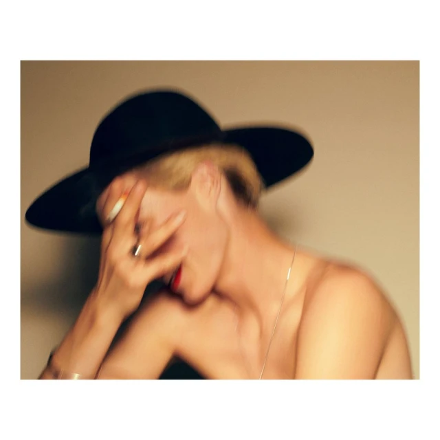 Abstract portrait of a woman with red lipstick wearing a large black hat, hand covering face, against a light tan background, blurred.
