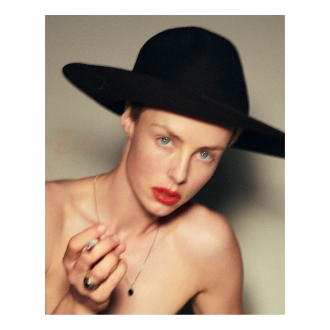 Close-up portrait of a fair-skinned person wearing a black wide-brimmed hat, red lipstick, and silver necklace against a neutral background.