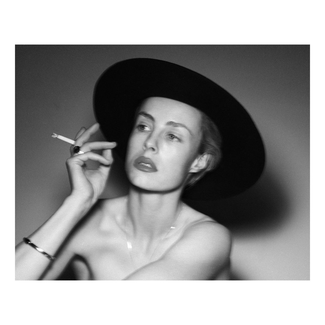 Monochrome portrait of a woman in a wide-brimmed black hat holding a lit cigarette, photographed by Theo Sion.