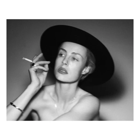 Monochrome portrait of a woman in a wide-brimmed black hat holding a lit cigarette, photographed by Theo Sion.