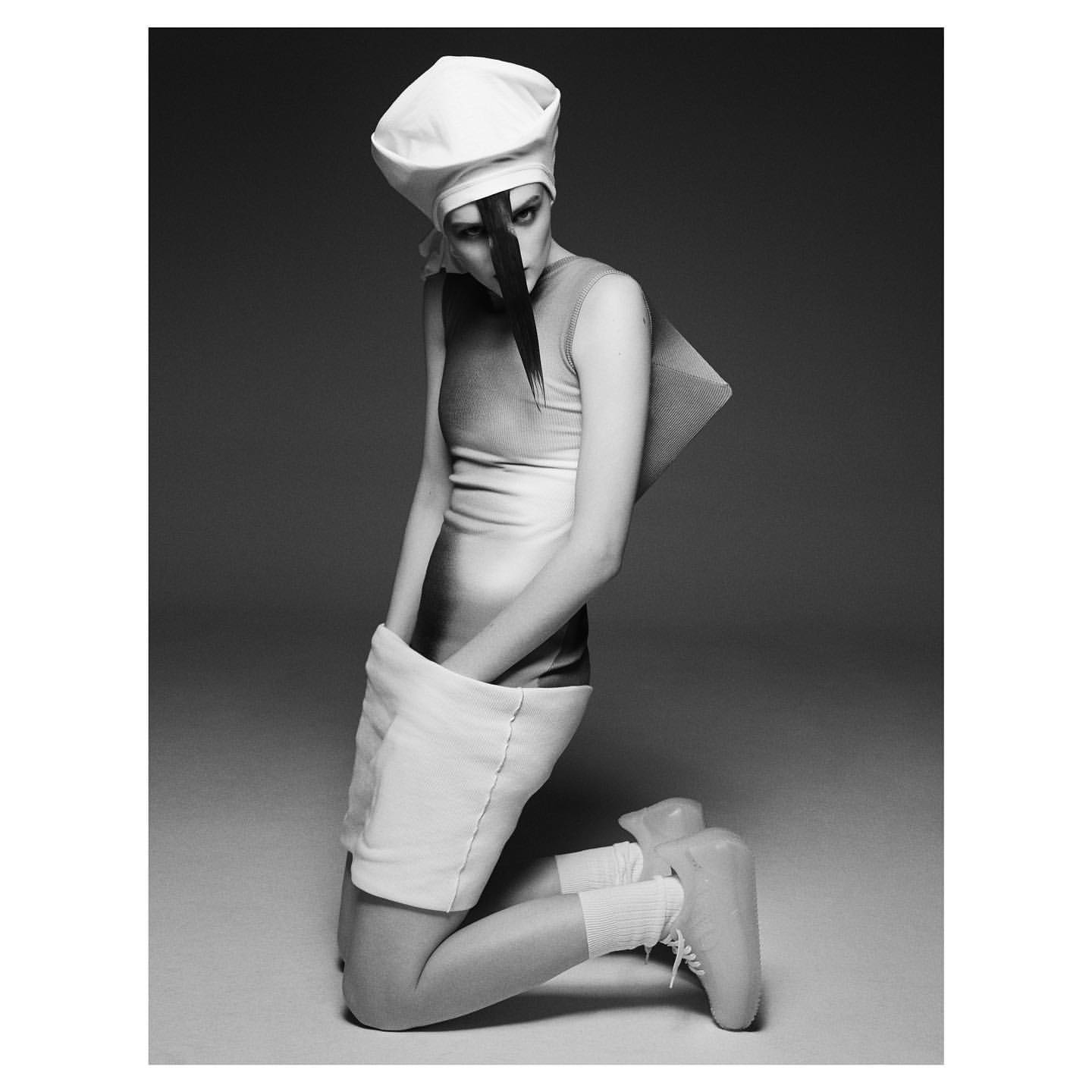 Monochrome fashion shot of a slender adult female model kneeling in a white hat, dress, socks, and sculptural platform shoes in a studio.