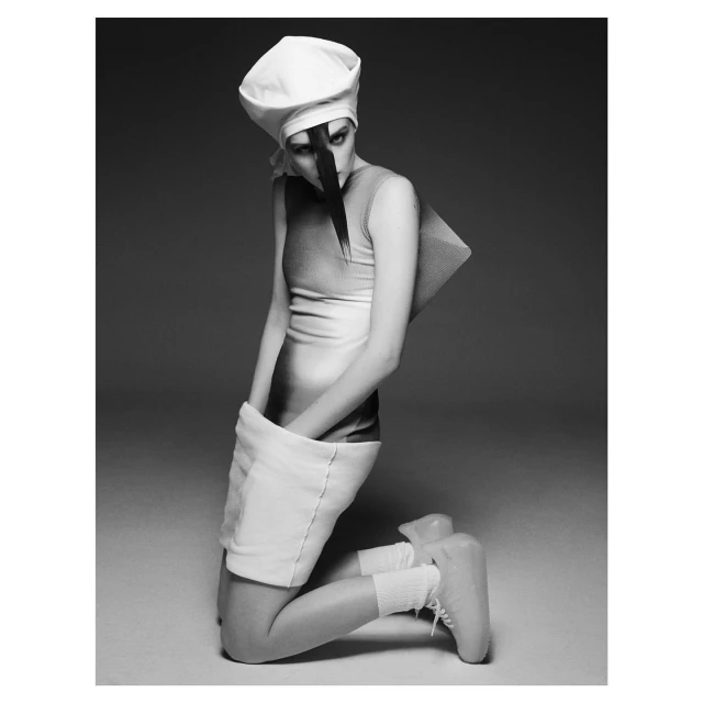 Monochrome fashion shot of a slender adult female model kneeling in a white hat, dress, socks, and sculptural platform shoes in a studio.