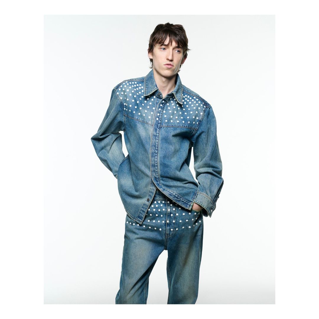 Male fashion model poses in a light denim studded shirt and jeans against a white background in a studio setting, full-length.