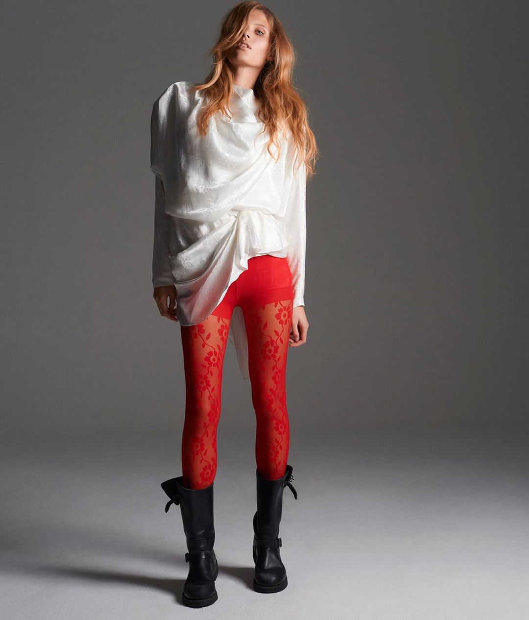 A long-haired, fair-skinned woman poses in a draped white top, patterned red leggings, and black boots against a neutral background.