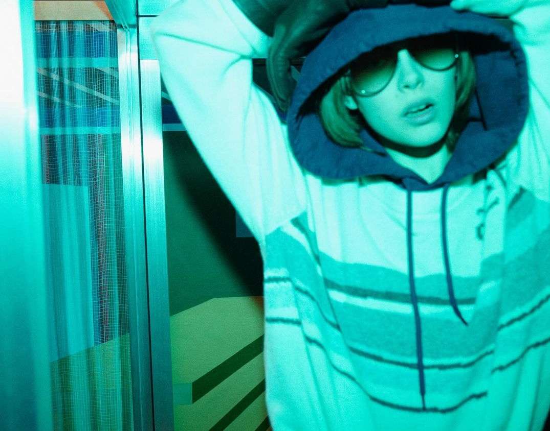 Teen poses in a hooded sweatshirt with sunglasses, illuminated by a blue and green neon light, creating a cool, youthful vibe.