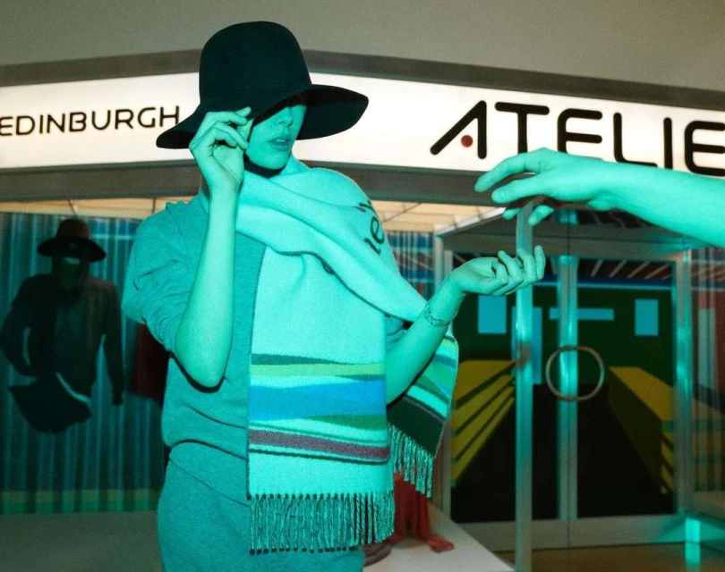 Woman in a gray outfit and hat poses in front of a store display with the word ATELIER in the background, bathed in teal lighting.
