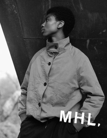 An androgynous-looking young adult in a khaki jacket poses in an outdoor urban setting with a textured dark background and partial natural light.