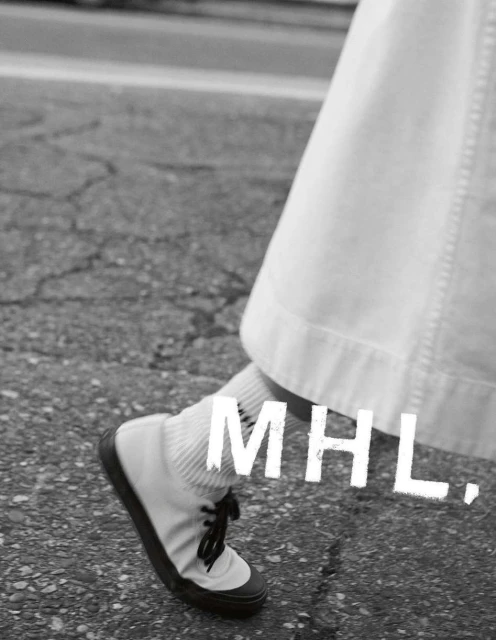 Close-up monochrome shot of a person walking in white sneakers and socks on cracked asphalt, featuring the MHL logo.