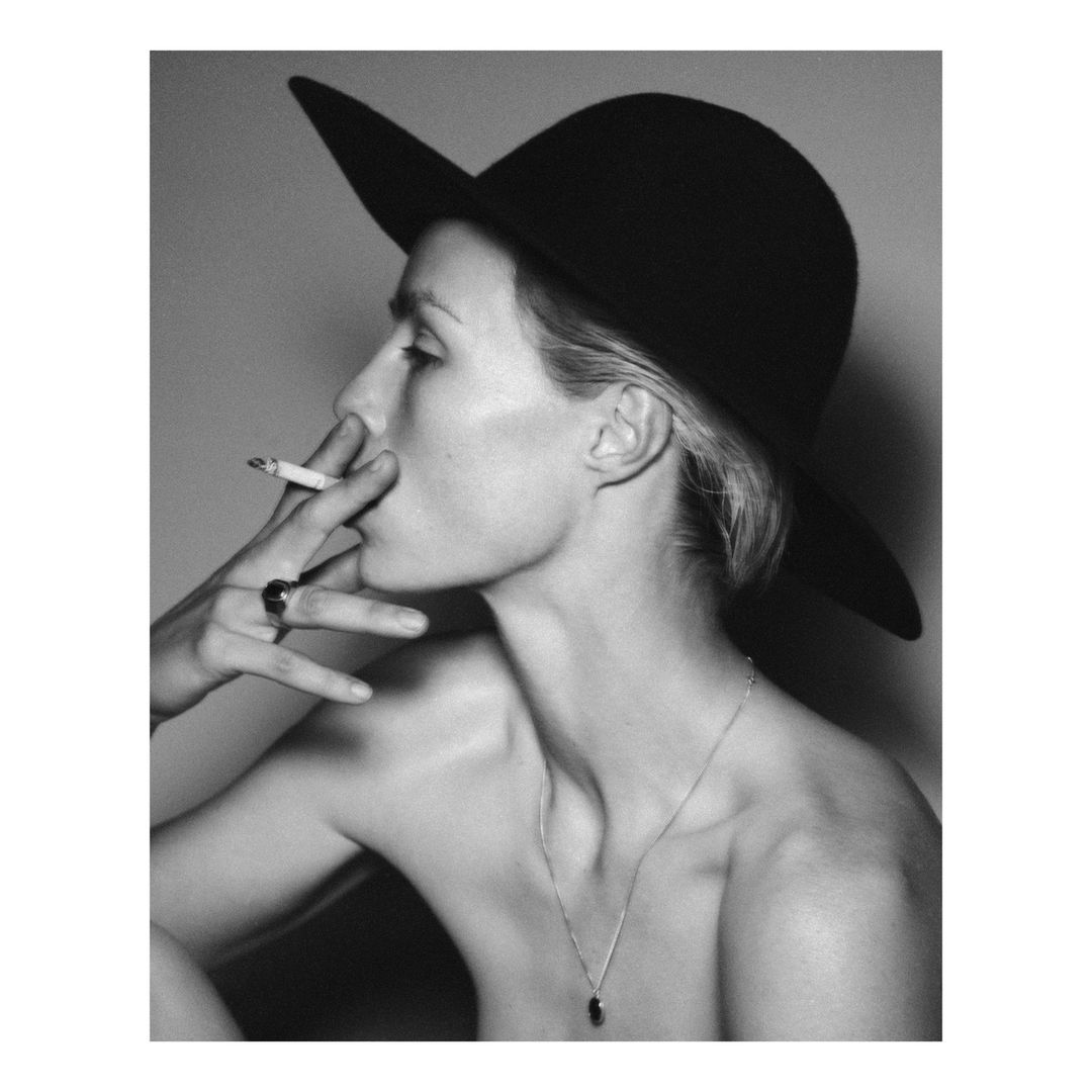 Black-and-white portrait of a fair-skinned adult woman in a hat smoking a cigarette, wearing jewelry, against a plain grey background.