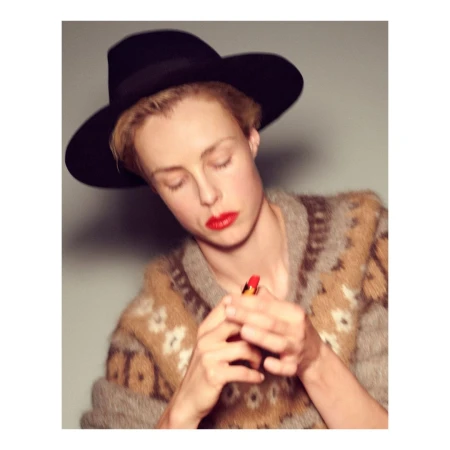 Model wearing a black fedora hat and patterned sweater holding a red lipstick tube, photographed by Theo Sion.