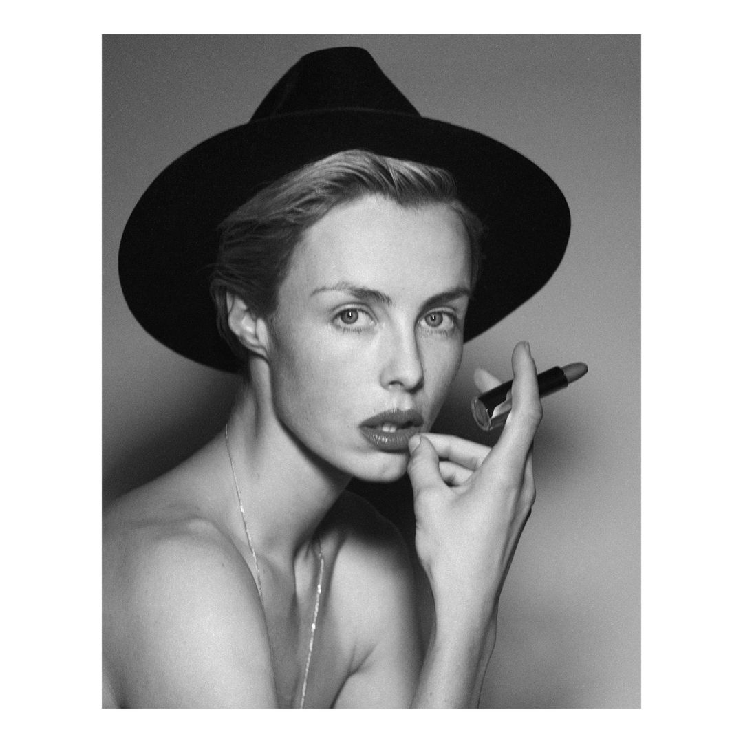 Black and white portrait of a woman wearing a wide-brim hat holding a lipstick tube, photographed by Theo Sion.
