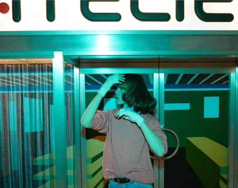 Person in pink sweater poses inside building with geometric mural, teal lighting, and text along ceiling in a medium shot.
