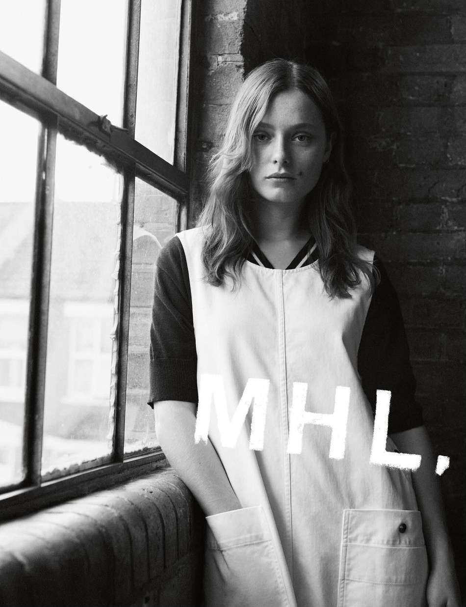 Black and white portrait of a young woman in a vintage-style overall dress, posing indoors near a window, with MHL text overlay.