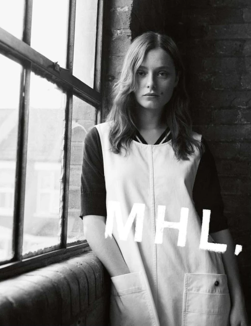 Black and white portrait of a young woman in a vintage-style overall dress, posing indoors near a window, with MHL text overlay.