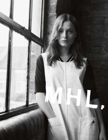 Black and white portrait of a young woman in a vintage-style overall dress, posing indoors near a window, with MHL text overlay.