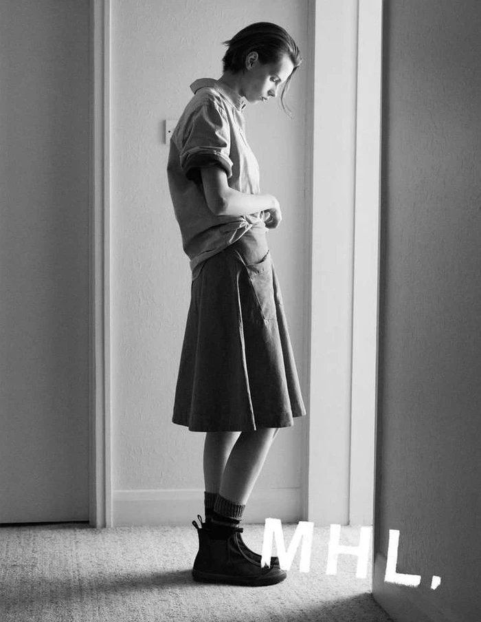 Monochrome full-length portrait of a woman standing in a doorway wearing a short-sleeve button-down shirt and skirt