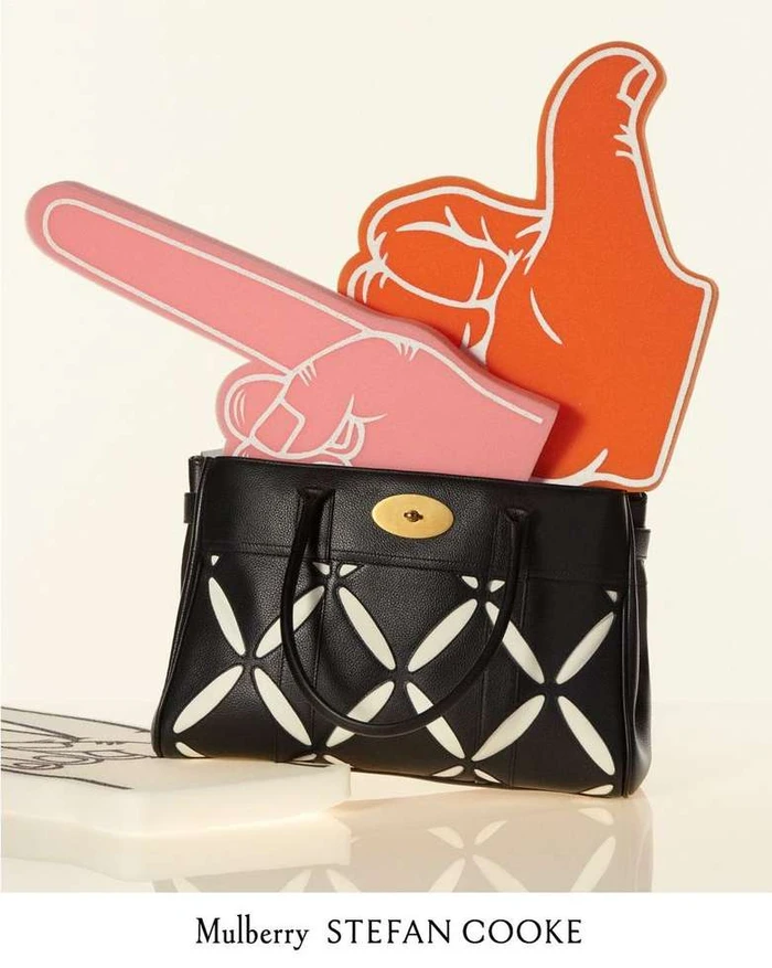 Black leather handbag with geometric cutouts holding pink and orange foam finger props against a white background.
