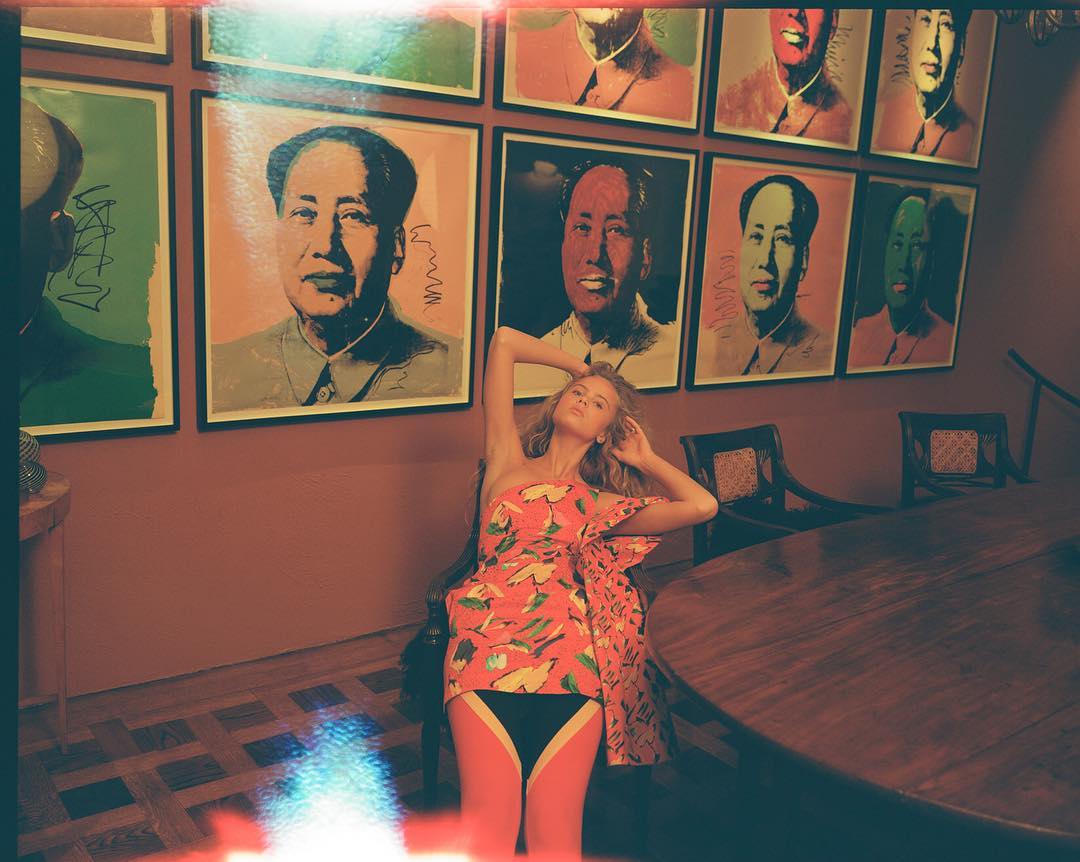 A woman in a patterned dress lounges in a chair in front of a wall adorned with framed portraits, next to a large table in an indoor setting.