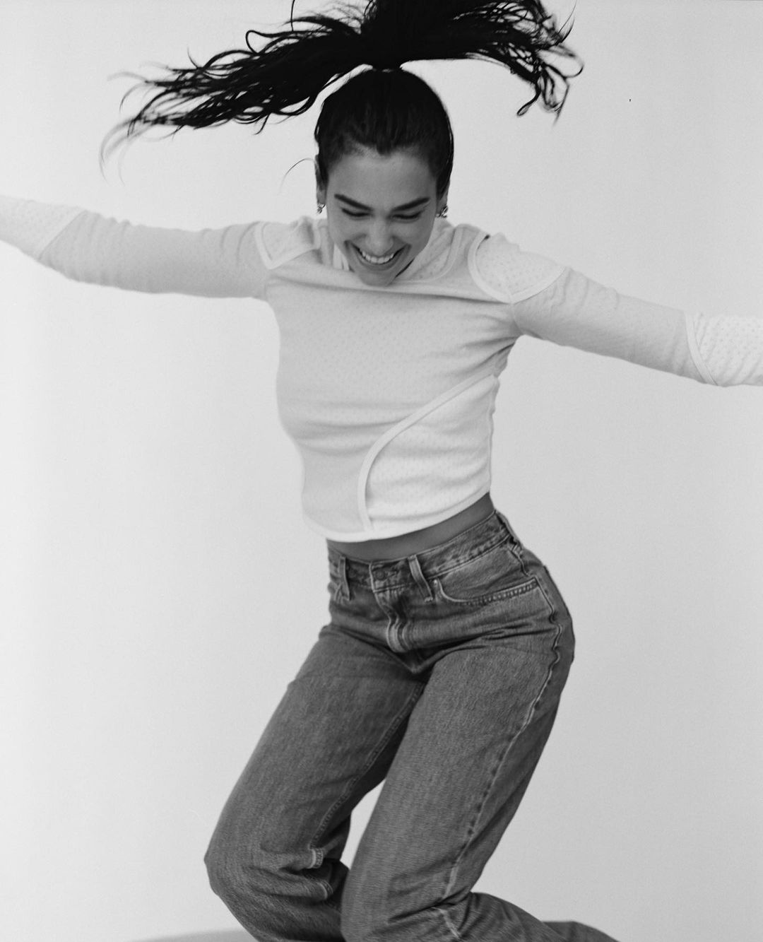 A woman jumps in a black-and-white studio shot wearing jeans and a long-sleeved shirt, smiling with her hair in a high ponytail.